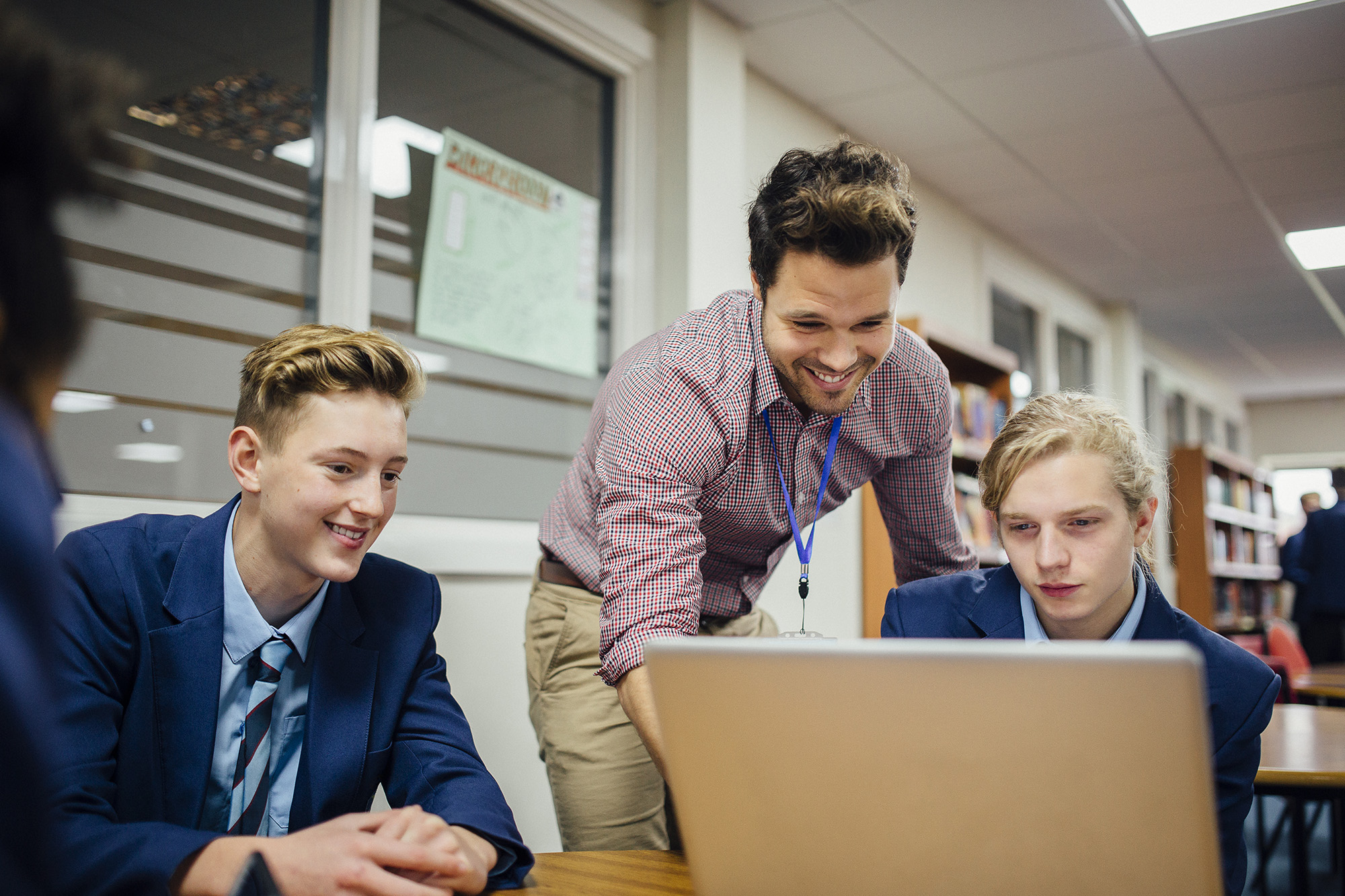 Teen students are working on a laptop in their school lesson. A teacher is leaning over them, helping them with some work.