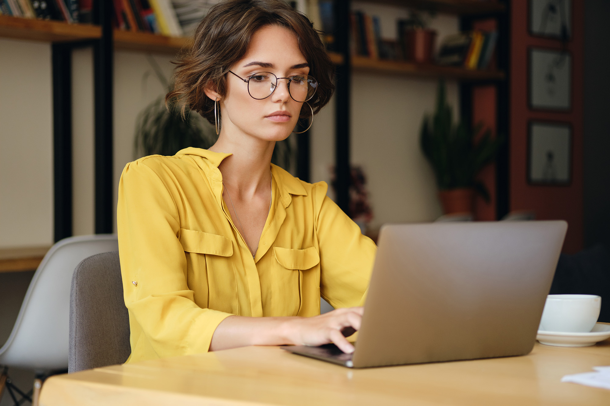 Young serious businesswoman in eyeglasses sitting at the desk thoughtfully working on laptop in modern office