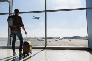 Young man is standing near window at the airport and watching plane before departure. He is standing and carrying luggage. Focus on his back